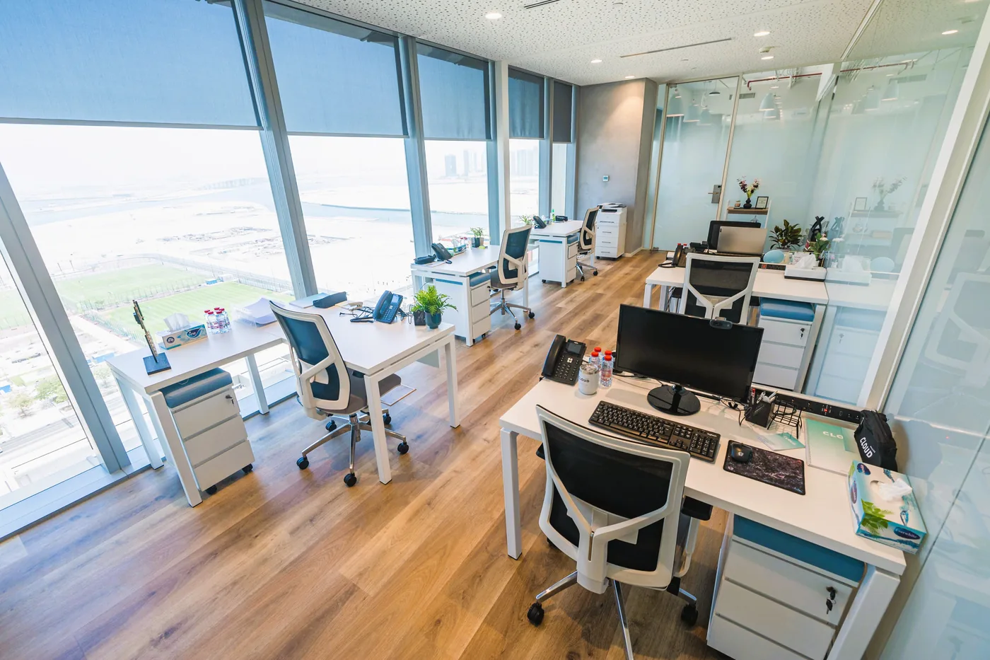 Desks along a wall of floor-to-ceiling windows, with blinds half drawn and a view over open ground beyond.