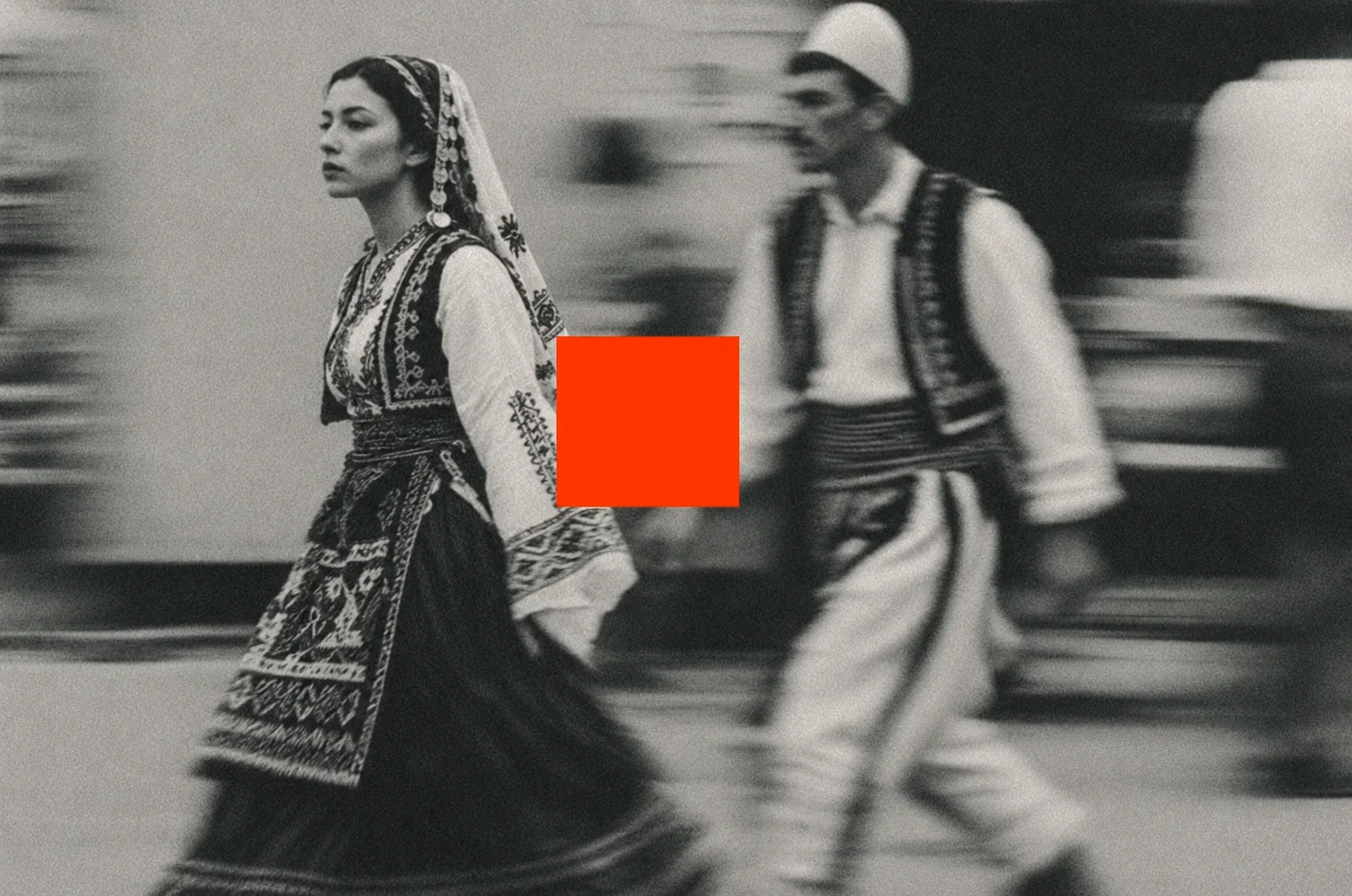 A woman and a man in traditional dress walking past, blurred by their movement, with a red square between them.