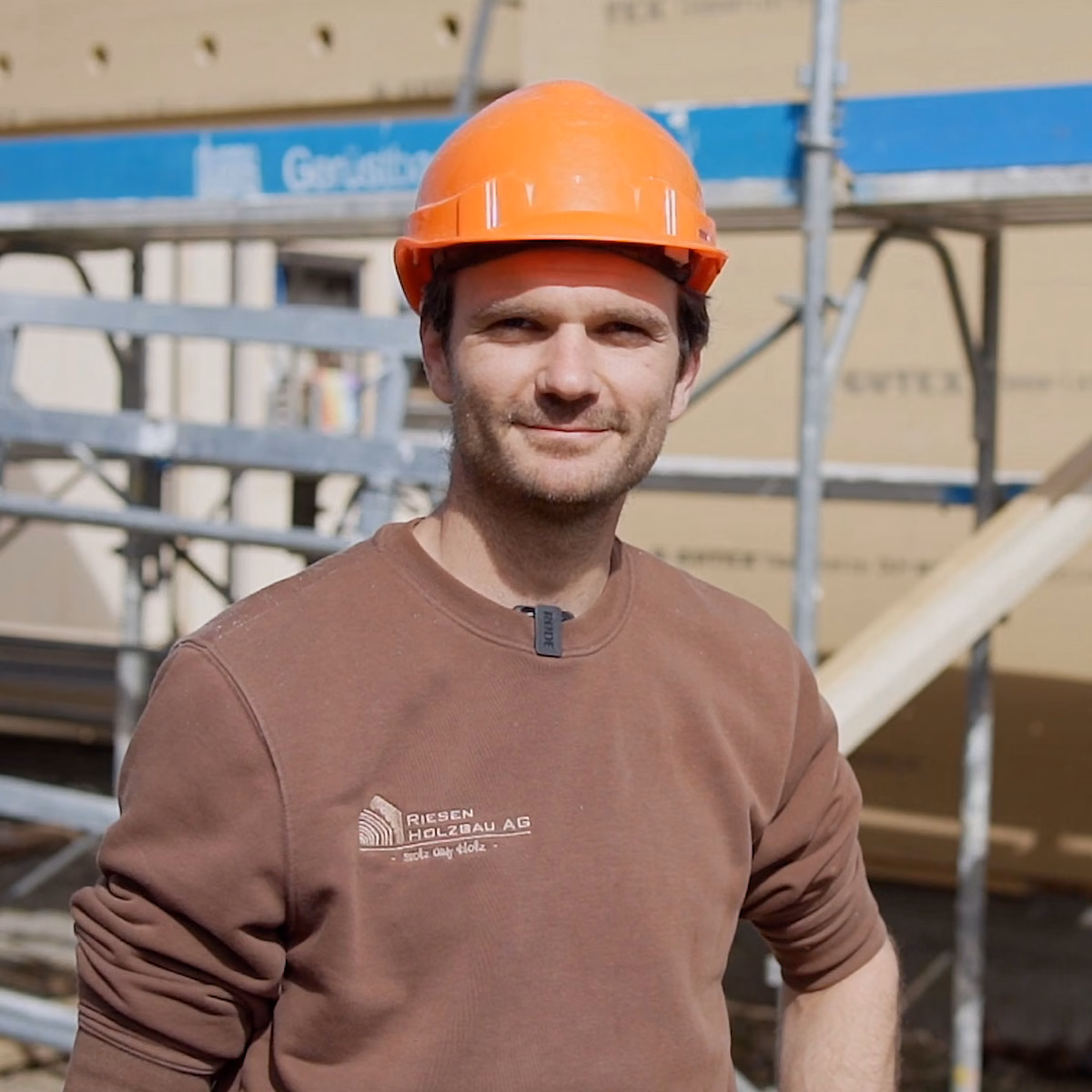 Riesen Holzbau AG carpenter in an orange helmet on site, the logo embroidered on the brown sweatshirt