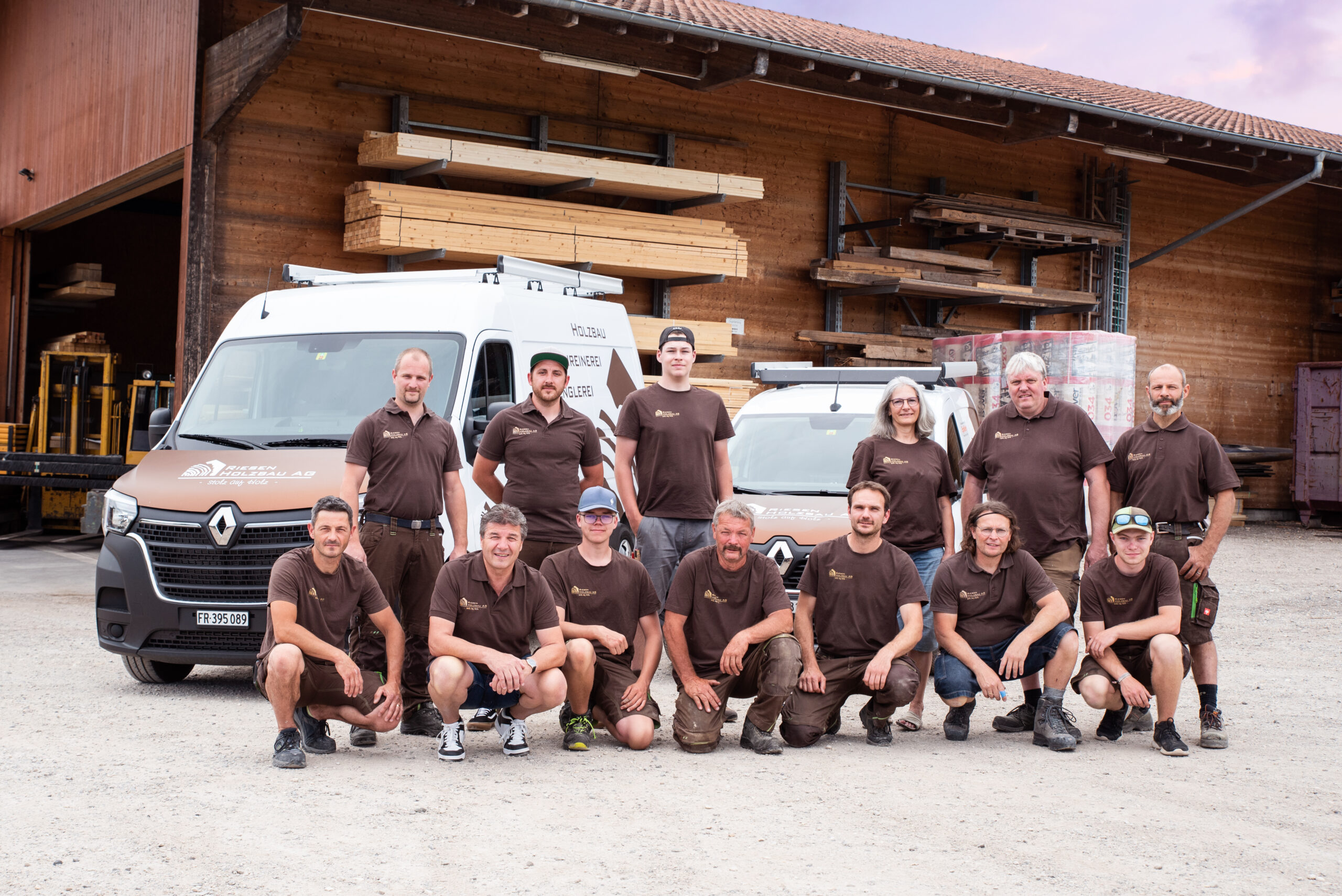 Riesen Holzbau AG team in brown branded workwear in front of two company vans and the timber store